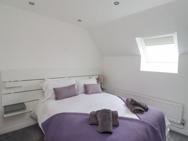 A bedroom with a double bed with purple pillows and blanket a window and a bedside lamp at Jara in Bamburgh