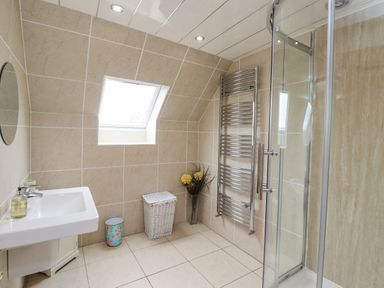 A bathroom with a wall-mounted sink a round mirror a small window a shower with glass door a towel radiator a wicker laundry basket a floral trash bin and a vase with flowers at Jara in Bamburgh