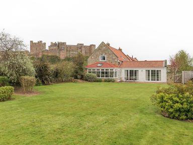 A house with a red tiled roof and stone walls with a large green lawn and bushes with a castle in the background at Jara Bamburgh