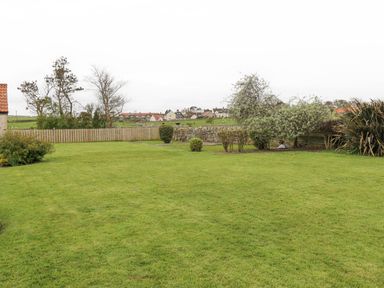 A garden with grass lawn shrubs trees a wooden fence and stone wall with houses in the background at Jara in Bamburgh