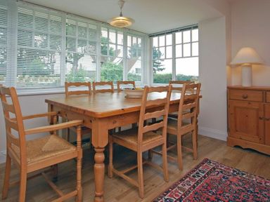 A dining room with a wooden table and six chairs near windows a wooden cupboard with a lamp and a rug on the floor at Stone Cottage in Bamburgh