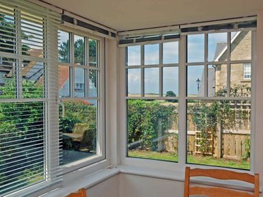 A corner of a room with large windows and blinds overlooking a garden and neighboring houses at Stone Cottage in Bamburgh