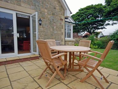 A stone patio with a round wooden table and four wooden chairs outside a stone house with an open door and garden in the background at Stone Cottage in Bamburgh