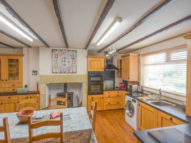 A kitchen with wooden cabinets a dining table a washing machine and a window with blinds at Creel Cottage Amble