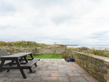 A patio with a wooden picnic table and bench overlooking a stone wall and grassy area with a view of the sea at Creel Cottage Amble