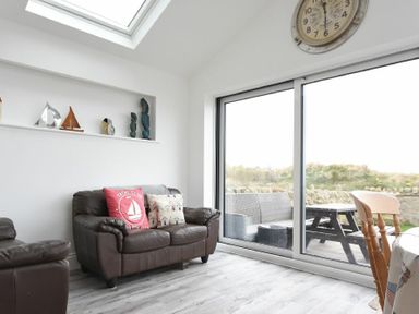 A living room with a brown leather sofa and armchair decorative sailboat ornaments a clock and sliding glass doors leading outside at Creel Cottage Amble