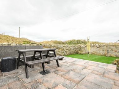 A patio with a picnic table and bench seating next to a low stone wall and grass area at Creel Cottage Amble