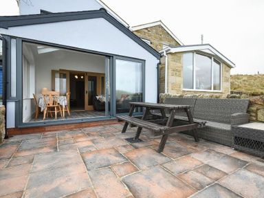 A patio with a wooden picnic table and rattan sofa outside a house with large glass doors at Creel Cottage Amble