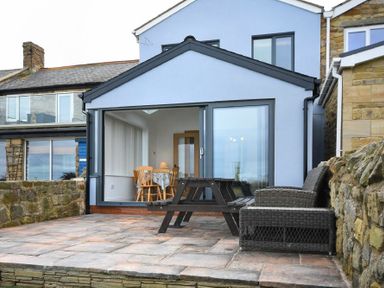 An outdoor patio area with a wooden picnic table and wicker chairs in front of a house with blue exterior walls and large sliding glass doors at Creel Cottage Amble