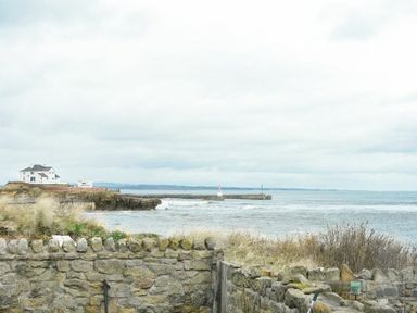 A coastal view with a stone wall in the foreground a house on a cliff and a pier extending into the sea at Creel Cottage Amble