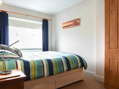 A bedroom with a bed covered in striped bedding a wooden nightstand with a lamp blue curtains and a wooden wardrobe at Creel Cottage Amble