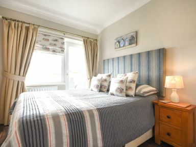 A bedroom with a striped bedspread and pillows a wooden nightstand with a lamp next to the bed and curtained windows at Creel Cottage Amble