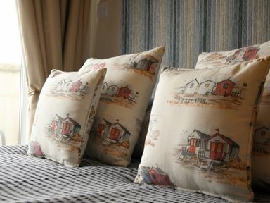 Decorative pillows with house prints on a striped bedspread near a window with curtains at Creel Cottage Amble
