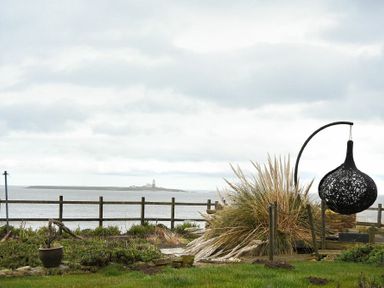 A garden with a black hanging chair and wooden fence overlooking the sea and an island with a lighthouse at Creel Cottage Amble