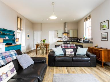 An open living room and kitchen with black leather sofas wooden dining table and blue cabinet at Baysyde in Alnmouth