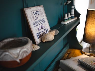 A shelf with a sign saying life is better at the beach seashells a small model lighthouse and boats next to a lamp on a wooden surface at Baysyde in Alnmouth
