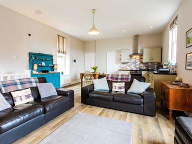 An open plan living room and kitchen with two black leather sofas a wooden dining table blue cabinet and kitchen appliances at Baysyde Alnmouth