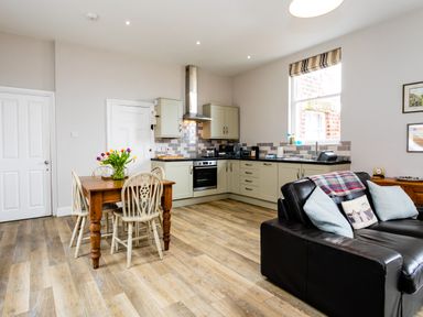 An open plan kitchen and living area with wooden dining table and chairs a black sofa and kitchen cabinets at Baysyde in Alnmouth