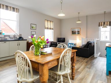 A kitchen and living area with a wooden dining table and chairs a vase with tulips a sitting area with black leather sofas and a television at Baysyde in Alnmouth