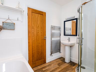 A bathroom with a wooden door a white sink with mirror a chrome towel rack a shower enclosure and a bathtub with a boat decoration on a shelf at Baysyde in Alnmouth