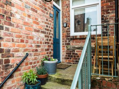 A brick porch with a blue door potted plants a window and a metal railing at Baysyde in Alnmouth