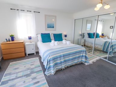 A bedroom with a double bed with striped blanket and blue pillows next to a wooden chest of drawers and mirrored wardrobe at Rathowen in Beadnell
