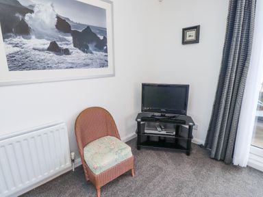 A corner of a room with a wicker chair with floral cushion a small TV on a black stand and ocean wave artwork on the wall at Rathowen Beadnell