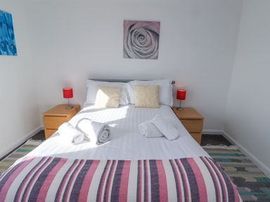 A bedroom with a bed with white linens towels and pillows two wooden nightstands with red lamps and wall art at Rathowen in Beadnell