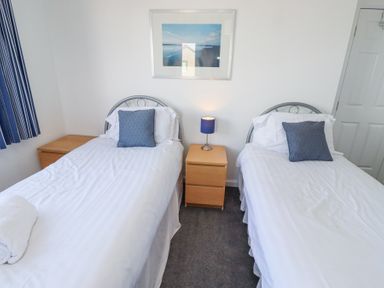 A bedroom with two single beds white bedding blue pillows a wooden nightstand with a lamp between the beds and a framed picture on the wall at Rathowen in Beadnell
