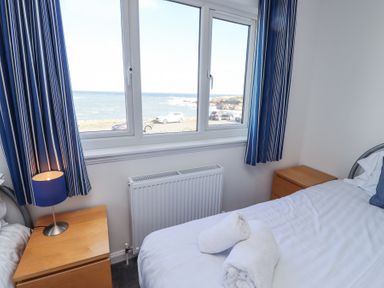 A bedroom with a bed showing rolled towels bedside tables with a lamp and window with blue curtains overlooking the sea at Rathowen in Beadnell
