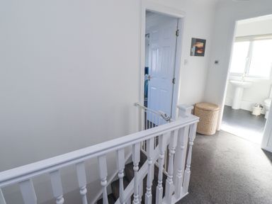 A hallway with a white staircase railing a child safety gate an open door leading to a bathroom a wicker basket and carpeted floor at Rathowen in Beadnell