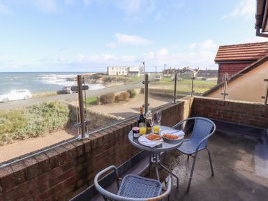 A balcony with a small round table and two chairs overlooking the sea with two croissants orange juice and beverages on the table at Rathowen in Beadnell