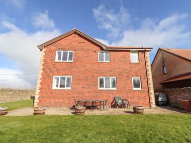 The back of a two-story brick house with six windows a patio area with chairs and a table and grass lawn at Rathowen in Beadnell