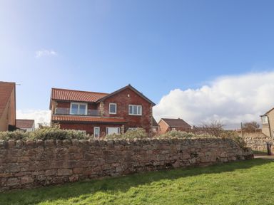 A red brick house with a tiled roof behind a stone wall and grassy area at Rathowen in Beadnell