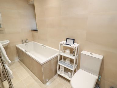 A bathroom with a bathtub a white toilet and a white shelving unit with toiletries at Nook Cottage in Embleton