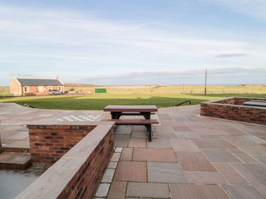 A patio with a wooden picnic table and benches overlooking a grassy field and a small stone building at Nook Cottage in Embleton