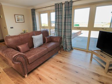 A living room with a brown leather sofa, blue plaid curtains, wooden floor, a TV on a wooden stand and large windows at Nook Cottage in Embleton