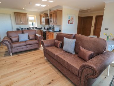 A living room with two brown sofas and a kitchen area with wooden cabinets at Nook Cottage in Embleton