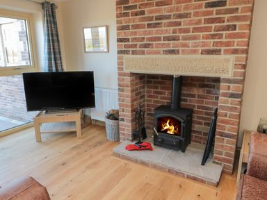 A room with a brick fireplace with a wood burning stove a TV on a wooden stand and a basket with firewood at Nook Cottage in Embleton