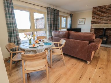 A dining area with a round table and wooden chairs next to a living room with a brown sofa and brick fireplace at Nook Cottage in Embleton