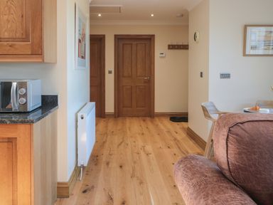 A hallway with wooden doors and flooring near a kitchen countertop with a microwave and a living area with a brown sofa and dining table at Nook Cottage in Embleton