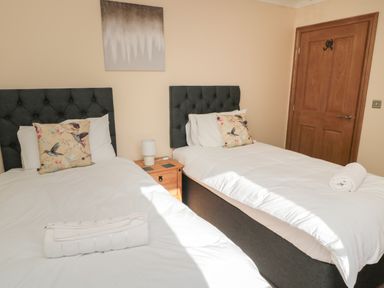 A bedroom with two single beds side by side with white bedding and floral pillows with a wooden door and bedside table with lamp at Nook Cottage in Embleton