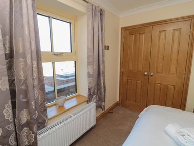 A bedroom corner with a window with patterned curtains a wooden closet and a bed with white linens at Nook Cottage in Embleton