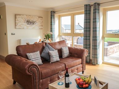 A brown leather sofa with cushions in a living room next to large windows with plaid curtains and a coffee table with wine and fruit at Byre Cottage in Embleton