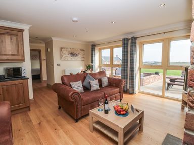 A living room with brown sofas a wooden coffee table with fruit and wine and large windows with curtains at Byre Cottage Embleton