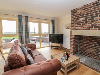 A living room with a brown sofa a wooden coffee table with wine glasses and fruit and a TV near a brick fireplace at Byre Cottage in Embleton