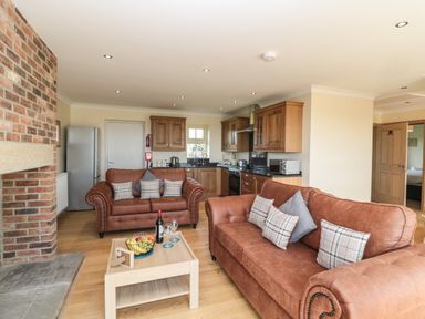 An open living room and kitchen area with brown sofas a wooden coffee table with fruit and wine and a brick fireplace at Byre Cottage in Embleton