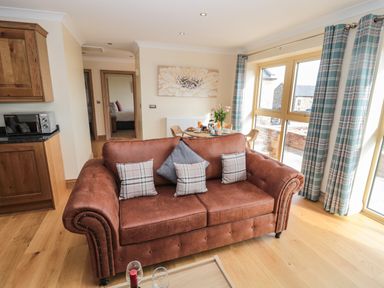 A living room with a brown sofa and cushions near large windows with checked curtains and a dining table with chairs at Byre Cottage in Embleton