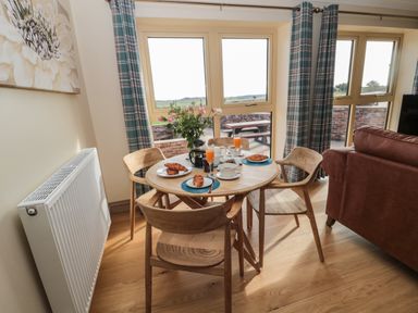 A dining area with a round wooden table set with croissants and drinks near windows with plaid curtains at Byre Cottage in Embleton