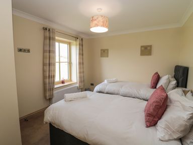 A bedroom with two single beds with white sheets and red pillows near a window with beige curtains at Byre Cottage in Embleton